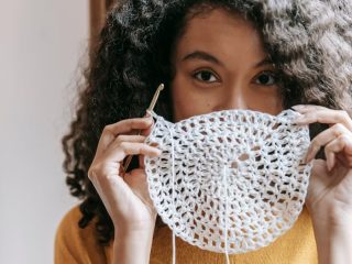 African American female with dark wavy hair wearing yellow pullover covering up mouth with white napkin with crochet and thread