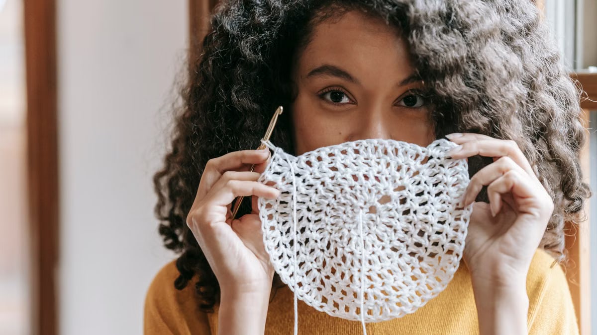 African American female with dark wavy hair wearing yellow pullover covering up mouth with white napkin with crochet and thread
