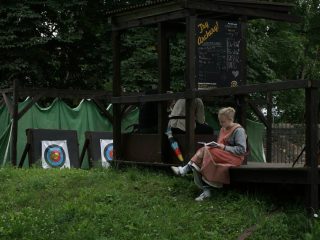 Young blonde woman wearing medieval clothing reading a book at an archery range