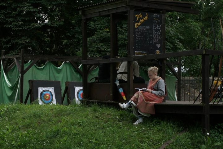 Young blonde woman wearing medieval clothing reading a book at an archery range