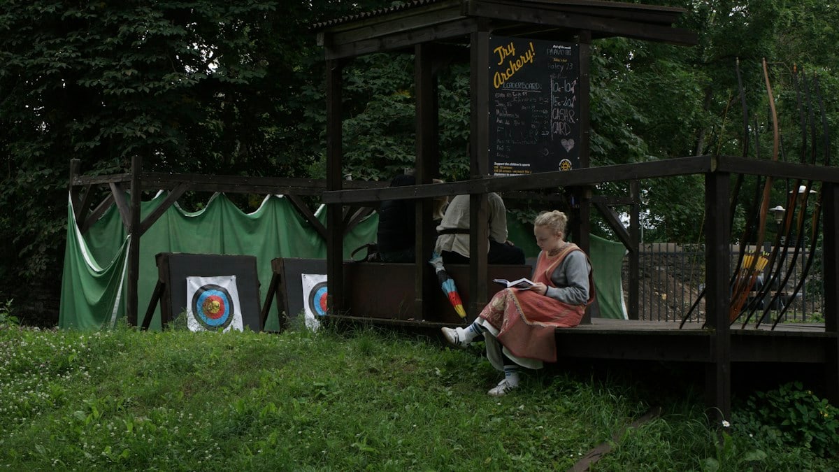 Young blonde woman wearing medieval clothing reading a book at an archery range