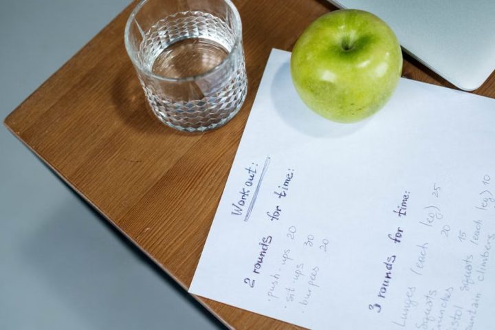 A green apple, glass of water, and workout plan on a wooden desk symbolize healthy living.